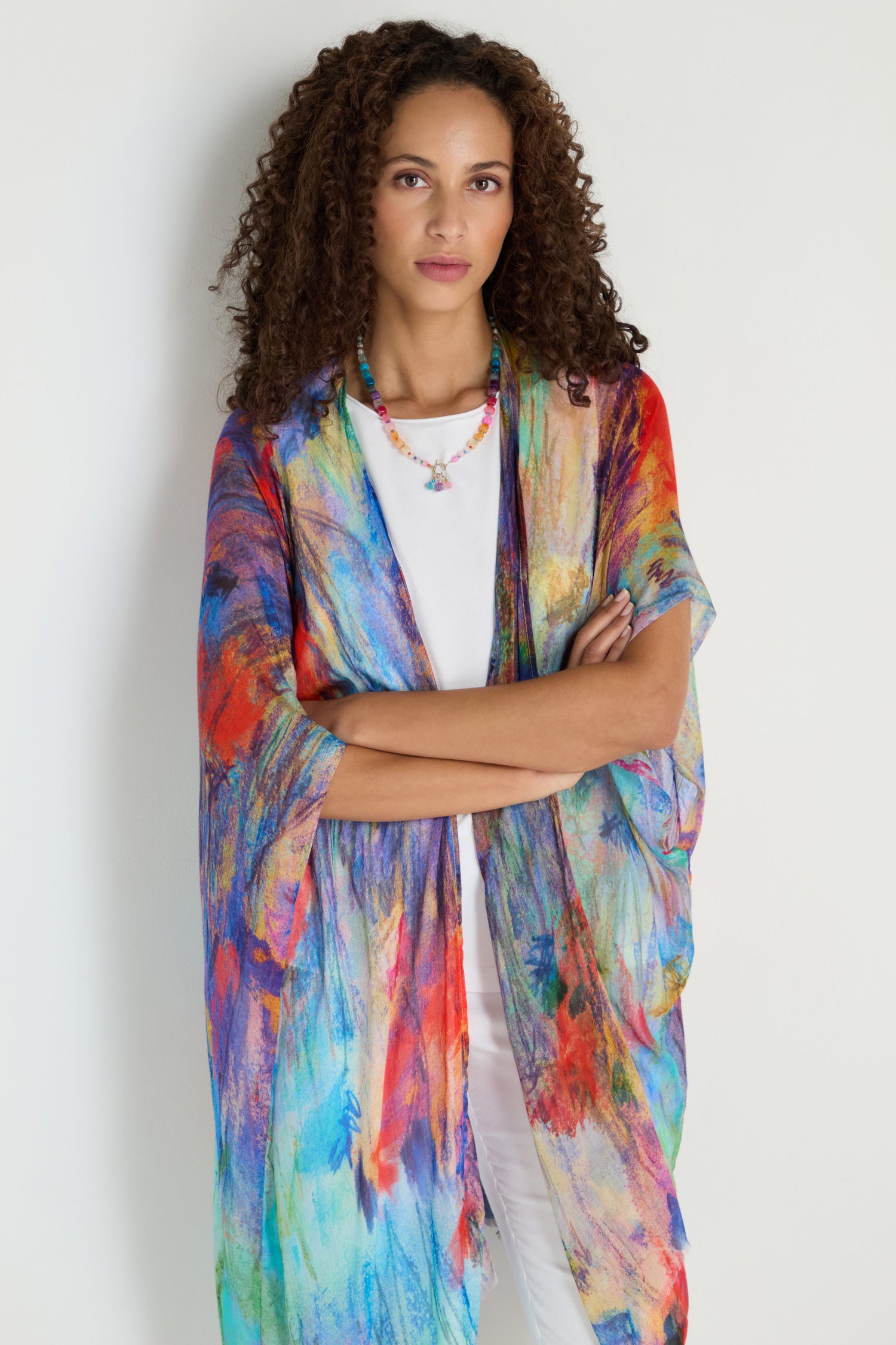 A woman with curly hair stands against a plain background, wearing the Soft Meadow Dreams Kimono over a white top and pants, arms crossed, complemented by a beaded necklace.