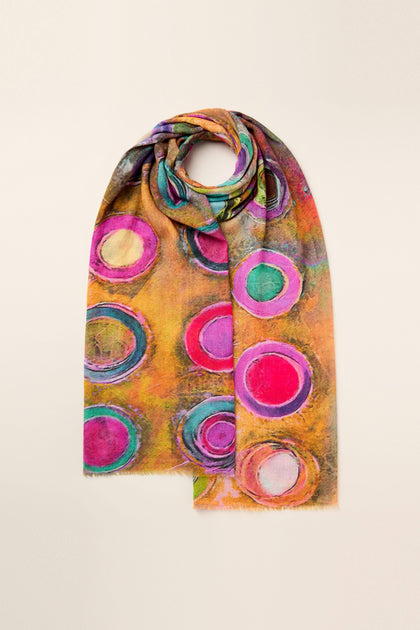 The Watercolour Circles Wool Mix Scarf features a vibrant pattern of overlapping circles in various shades of pink, green, and blue on a warm-toned background. It is neatly folded and displayed against a plain backdrop.