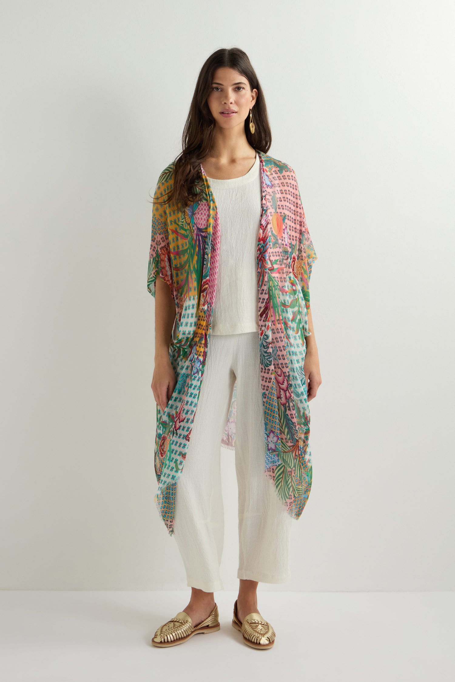 A woman in a vibrant Botanical Patchwork Kimono over a white outfit stands against a plain background, paired with gold sandals.