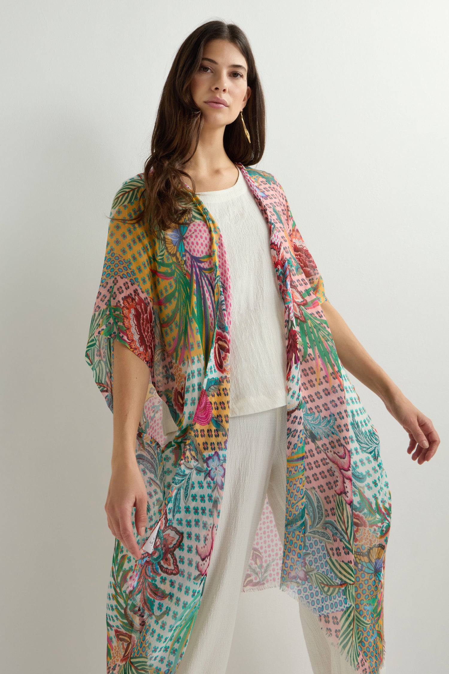 Against a plain backdrop, an individual elegantly wears the Botanical Patchwork Kimono, a lightweight and versatile garment with floral prints, over a pristine white outfit.