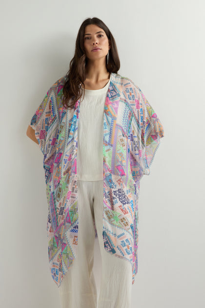 A woman poses against a plain backdrop, elegantly donning the Soft Graphic Patchwork Kimono over a white outfit; its vibrant design captivates as she looks forward with hands in pockets.