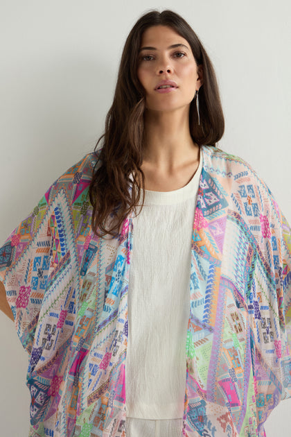 A woman elegantly stands against a plain background, draped in the Soft Graphic Patchwork Kimono over a white outfit.