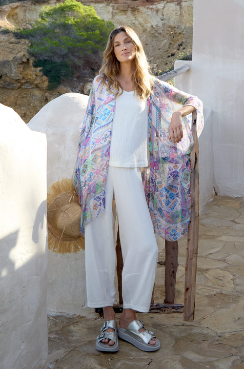 A woman in a white outfit and the "Modern Mosaic Kimono" stands by a rustic railing outdoors, accompanied by a straw hat. The stone walkway and lush greenery create a serene backdrop for her colorful geometric ensemble.