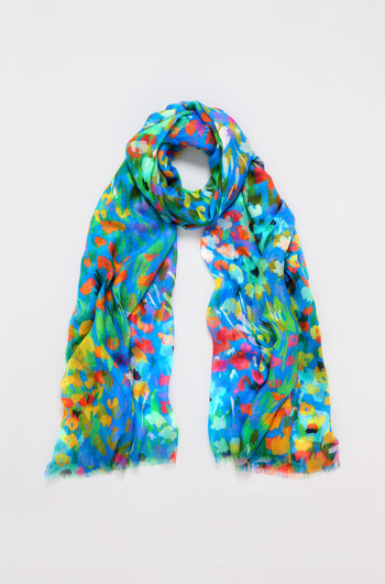 The Abstract Painted Garden Scarf features a vibrant mix of blue, green, orange, and yellow brushstrokes forming a floral-like abstract design. It appears lightweight and airy as it sits loosely coiled on a plain white background.