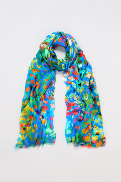 The Abstract Painted Garden Scarf features a vibrant mix of blue, green, orange, and yellow brushstrokes forming a floral-like abstract design. It appears lightweight and airy as it sits loosely coiled on a plain white background.