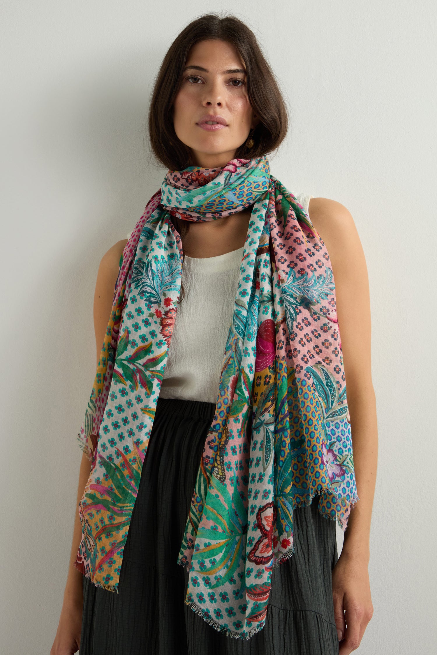 A woman effortlessly showcases her style, wearing a vibrant floral Botanical Patchwork Scarf over a white sleeveless top and dark skirt against a plain backdrop.