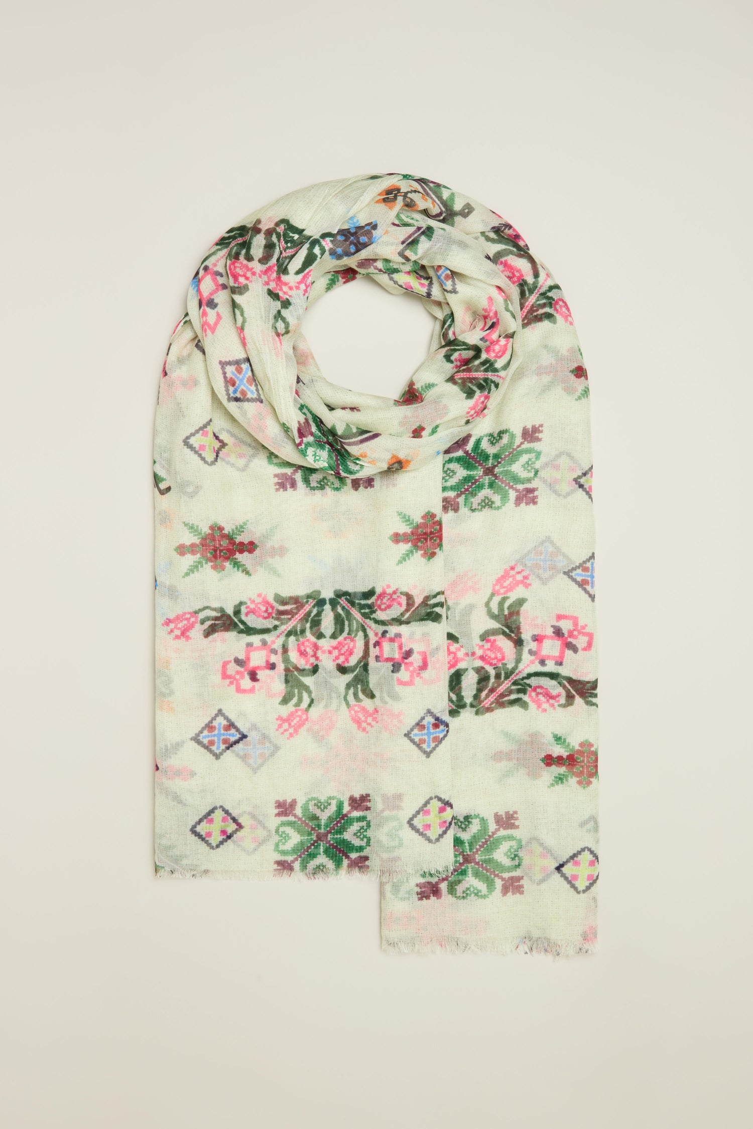 The Cross Stitch Print Scarf, folded and embellished with pink, green, and blue floral and geometric motifs, elegantly rests on a simple surface, highlighting its intricate design against a light backdrop.