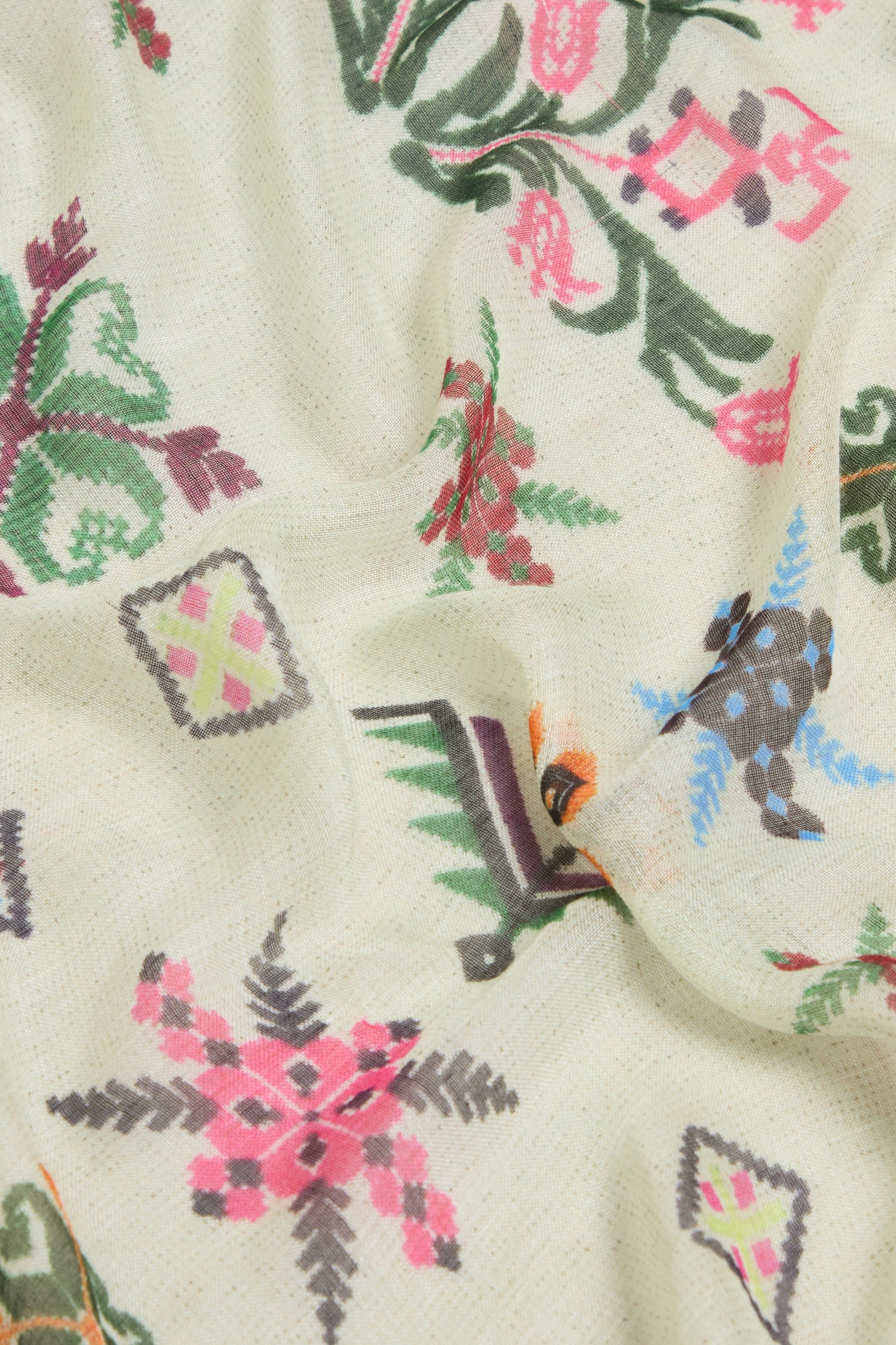 The Cross Stitch Print Scarf features a cream background with vibrant floral and geometric motifs in pink, green, blue, and black. Intertwining star shapes and floral designs evoke traditional embroidery styles.