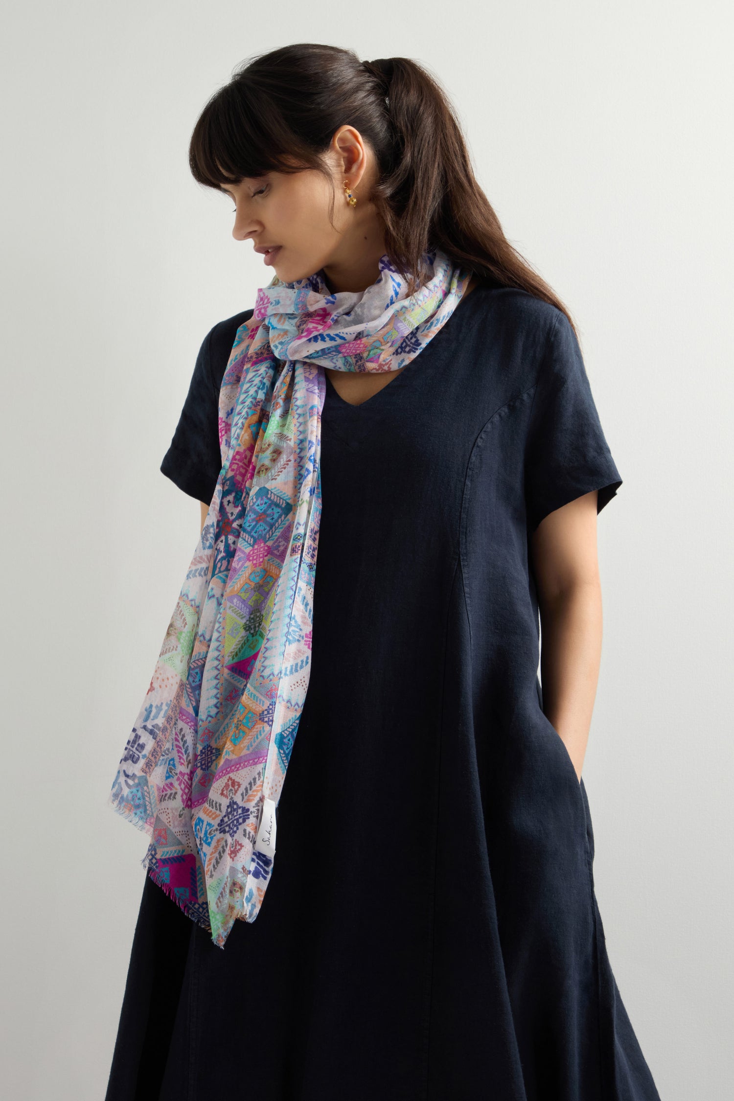 A person in a dark dress with hands in pockets, looking to the side. The Soft Graphic Patchwork Scarf with geometric and abstract prints is draped around their neck, adding a versatile and timeless touch of style.