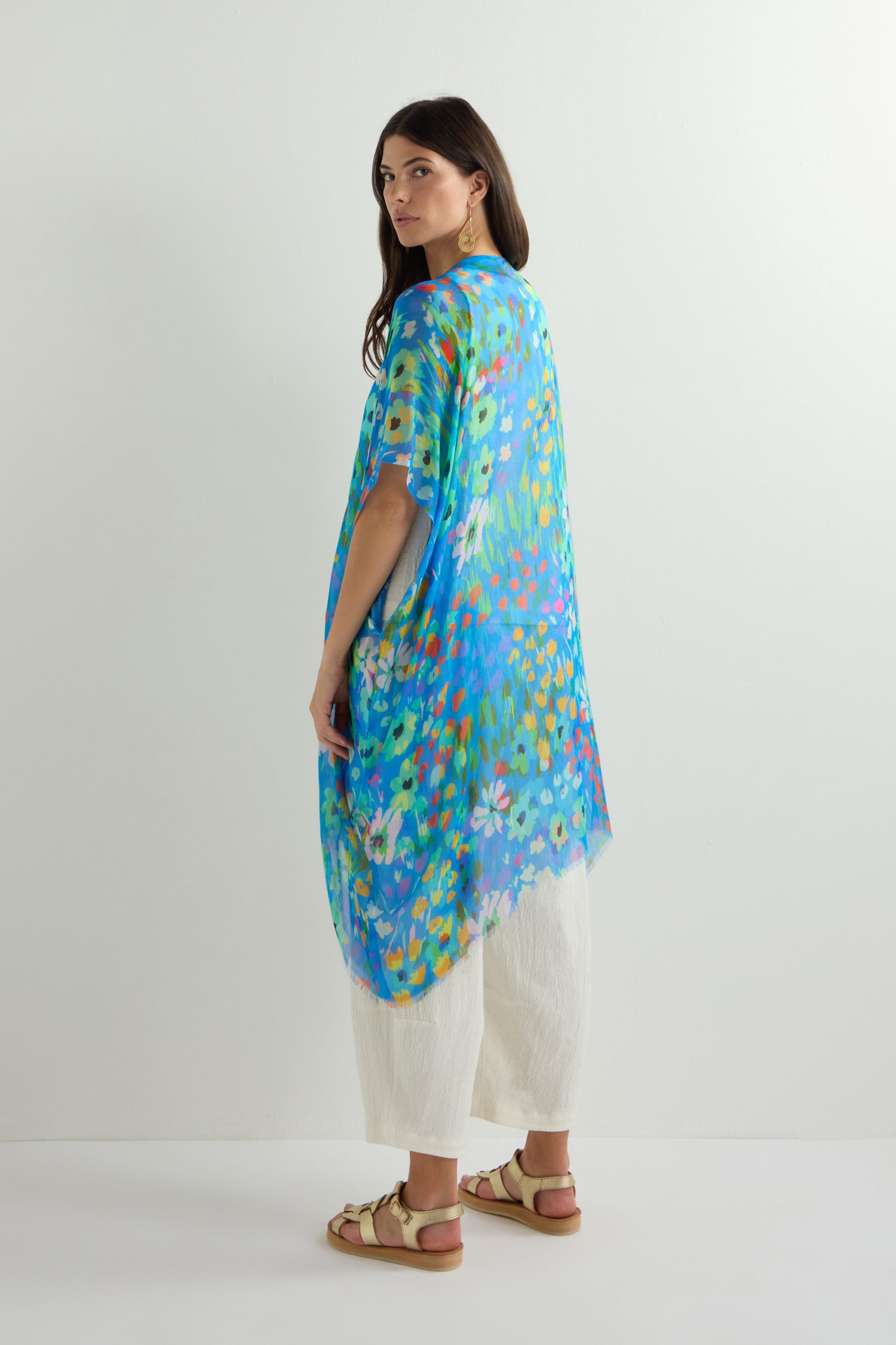 A woman stands against a plain backdrop wearing the Abstract Painted Garden Kimono, its blue floral design and lightweight open-front silhouette elegantly draping over white pants and sandals.