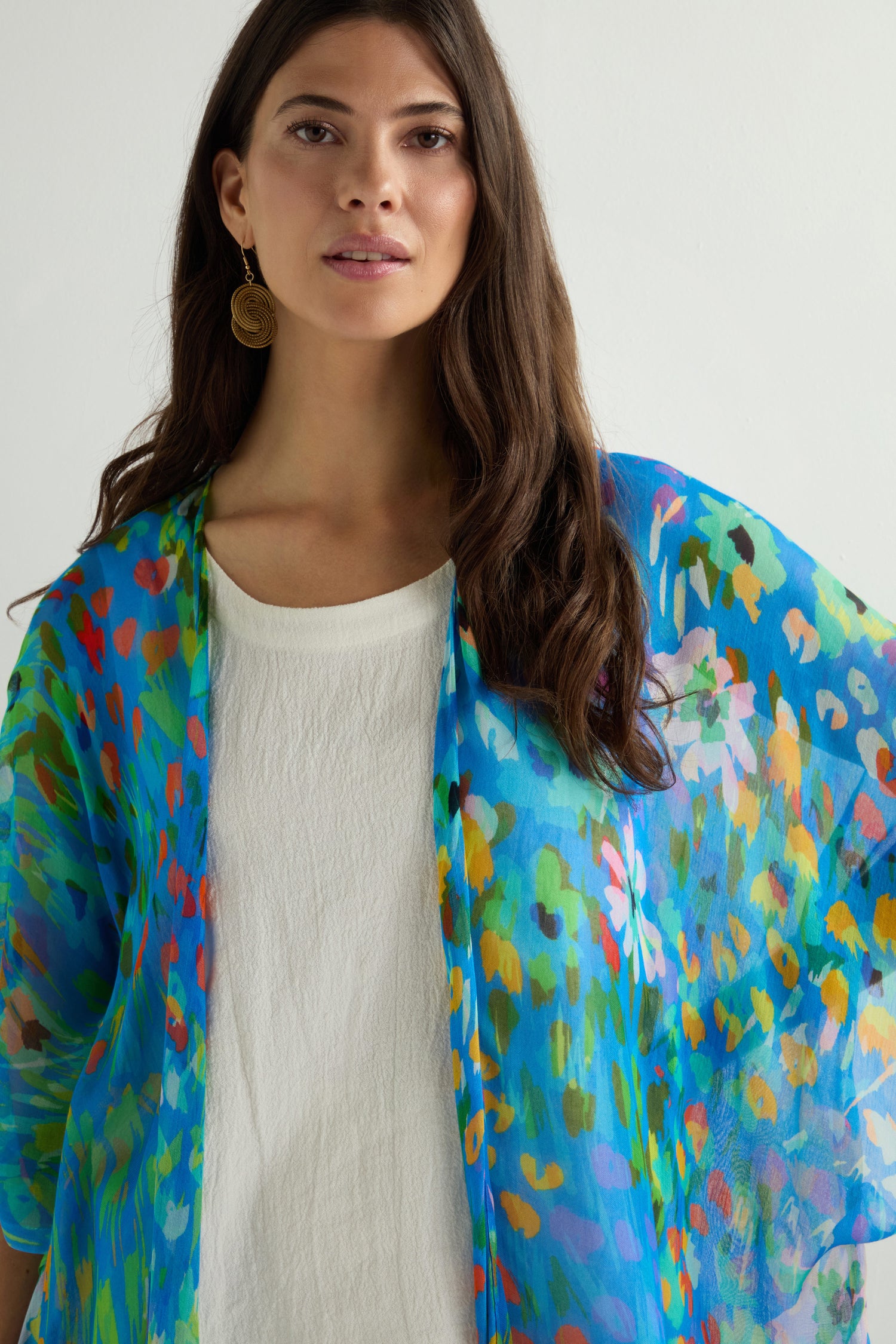 A person in a white top accessorizes with the Abstract Painted Garden Kimono, featuring a colorful blue pattern and flowy open-front design. Against a plain backdrop, their long hair and a gold circular earring are visible.