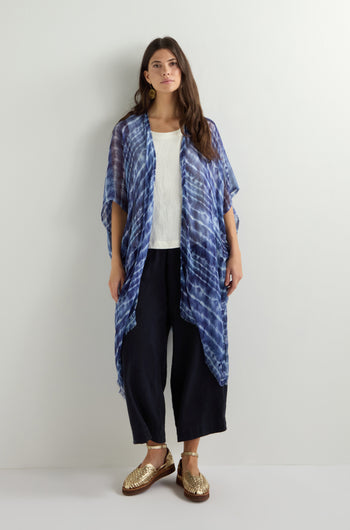 A person stands against a neutral background wearing the Tie Dye Print Kimono in blue over a white top and dark pants, paired with woven shoes. The open-front design enhances the lightweight cover-up's relaxed elegance.