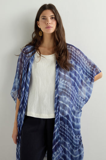 A woman in a Tie Dye Print Kimono with an open-front design stands against a plain light background, pairing the elegant piece with a white top and black pants for a chic and effortless look.