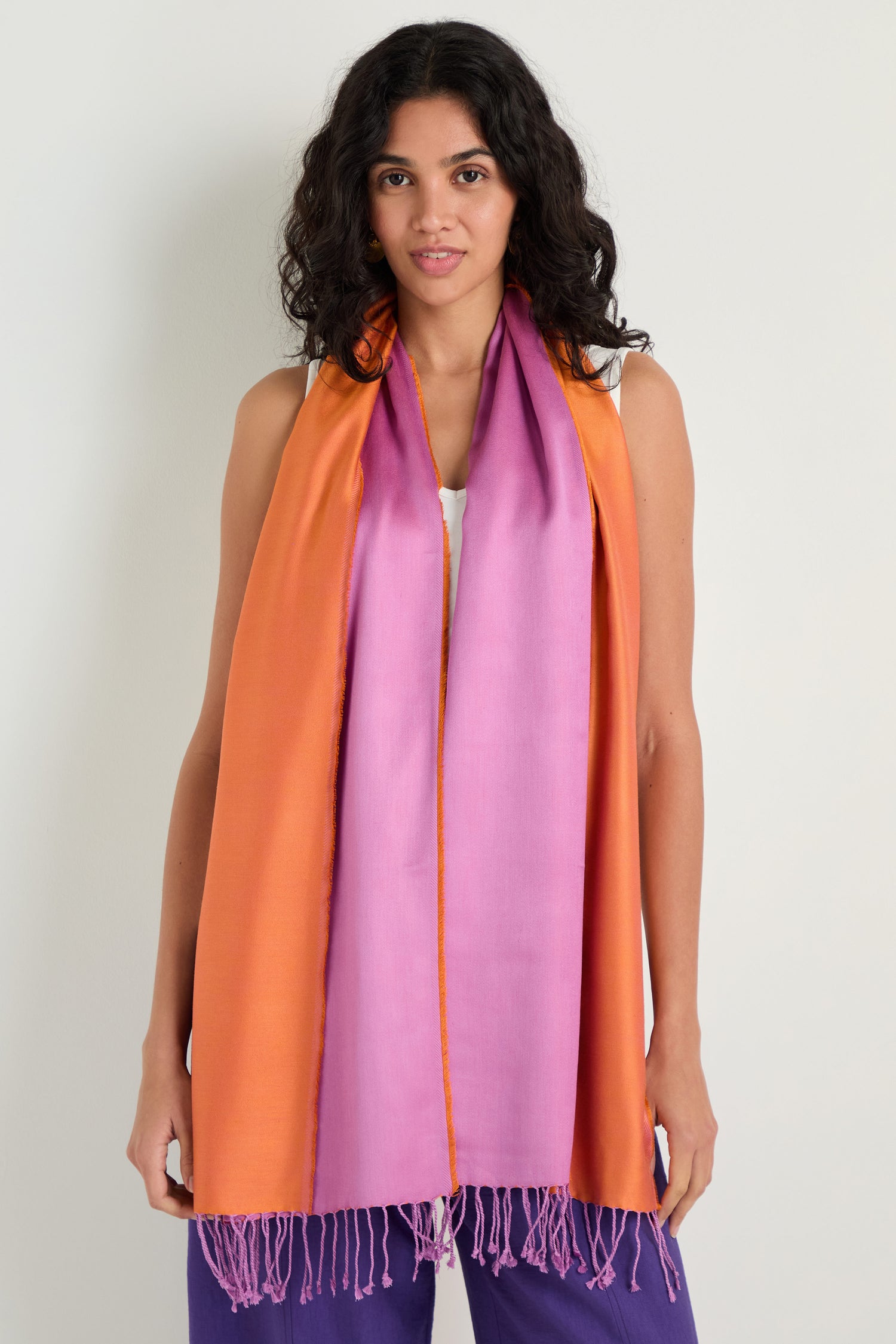 A woman in a white top and purple pants wears the Two Tone Silk Scarf, featuring colourblocking, fringed ends, and a luxurious sheen, draped around her neck against a plain light background.