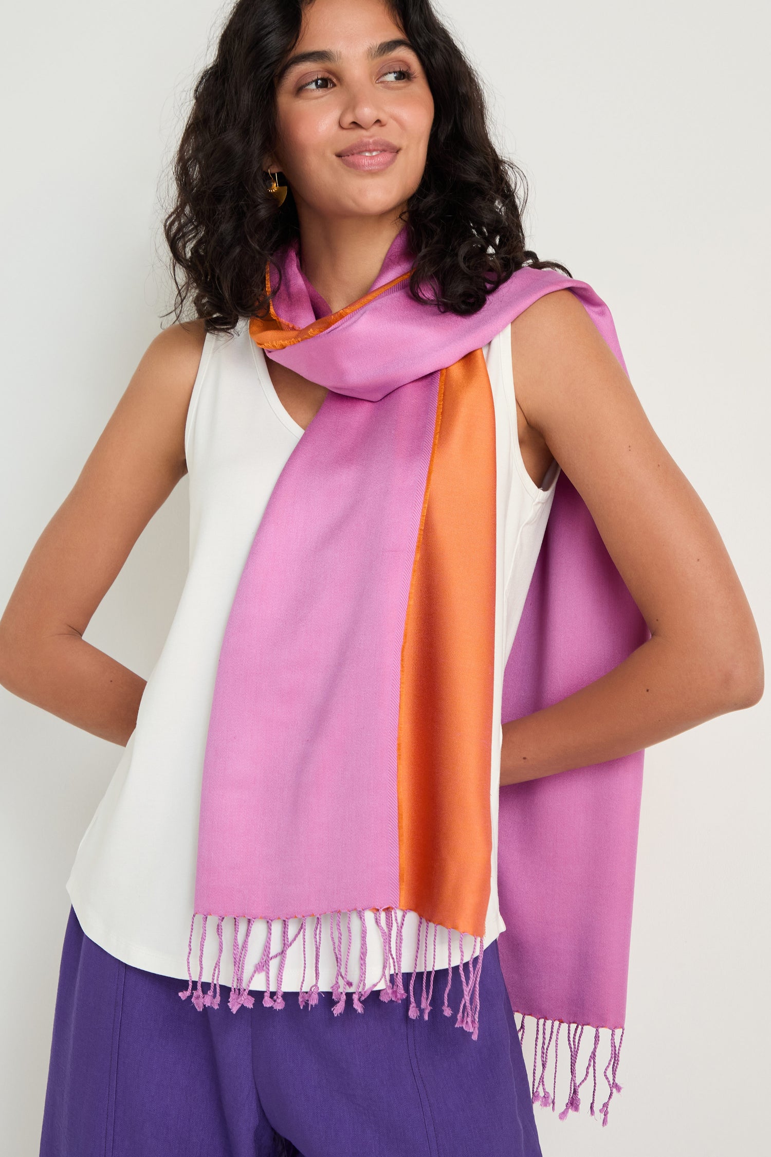 A woman stands against a plain background, wearing a sleeveless white top, purple pants, and the Two Tone Silk Scarf with a luxurious sheen draped around her neck and shoulder.