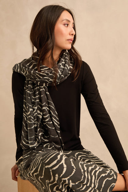 A woman sits on a wooden stool, wearing a black long-sleeve shirt and the Dancing Waves Scarf—a lightweight winter accessory featuring a striking wave pattern—paired with a matching skirt, looking to the side against a beige background.