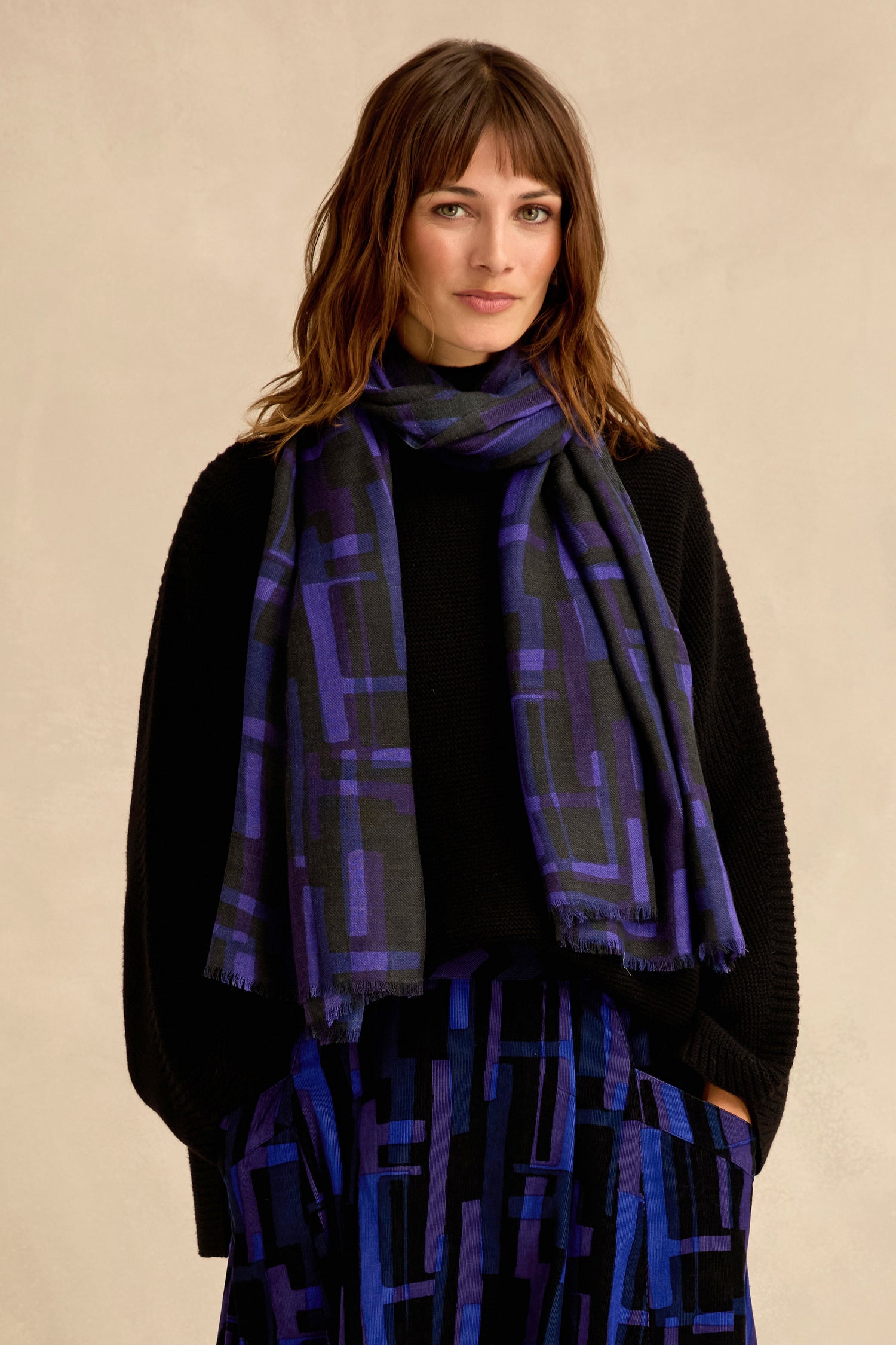 A woman with shoulder-length brown hair and bangs wears a black sweater, a layered geometric print skirt, and the Layered Geometric Wool Scarf in purple and black against a beige background.