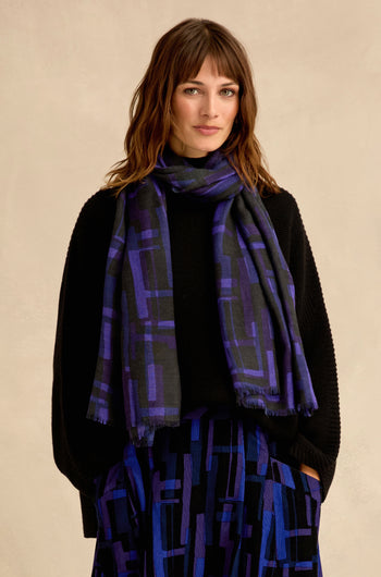 A woman with shoulder-length brown hair and bangs wears a black sweater, a layered geometric print skirt, and the Layered Geometric Wool Scarf in purple and black against a beige background.
