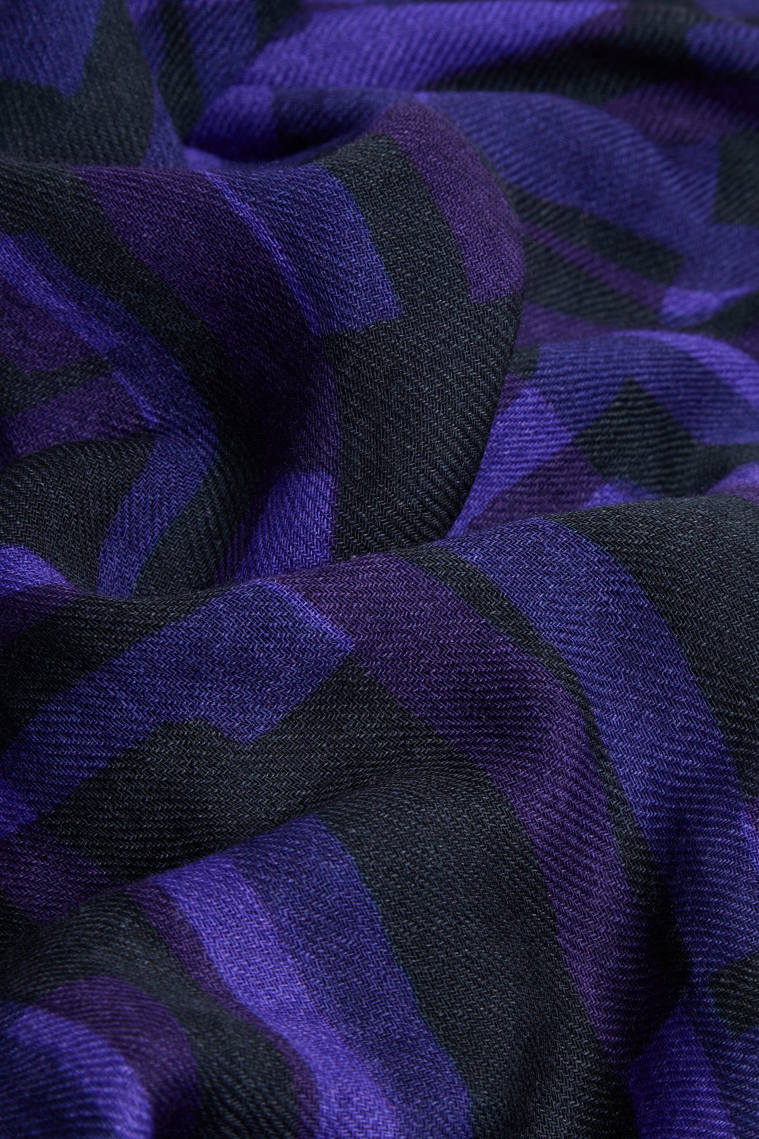 A close-up of the Layered Geometric Wool Scarf displays its purple and black geometric print, highlighting the fabric’s folds and texture.