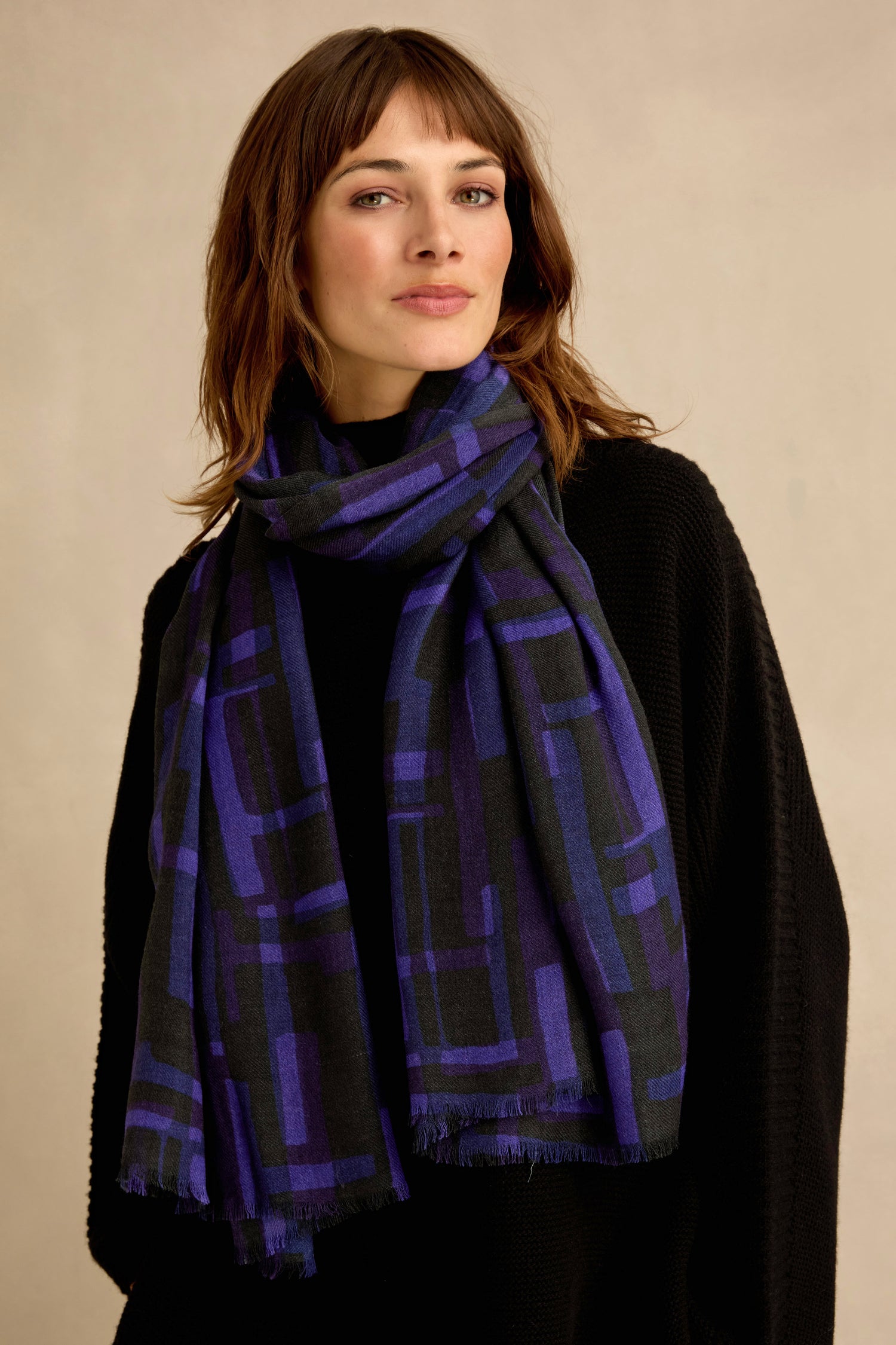 A woman stands against a plain beige background wearing a black sweater and the Layered Geometric Wool Scarf in blue and black.