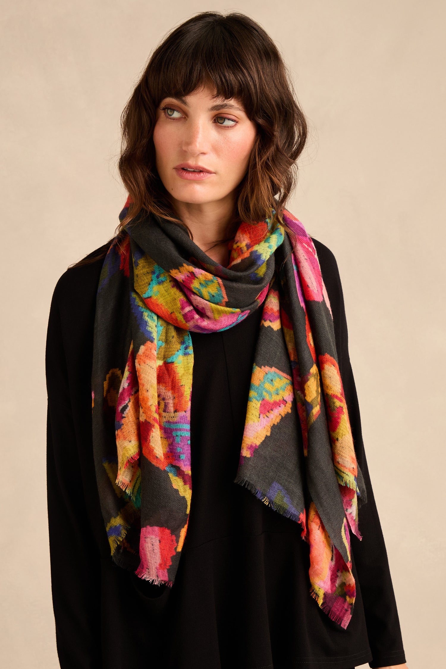 A woman stands against a plain beige background, looking slightly to the side, wearing a black top and the Pixelated Floral Wool Scarf.