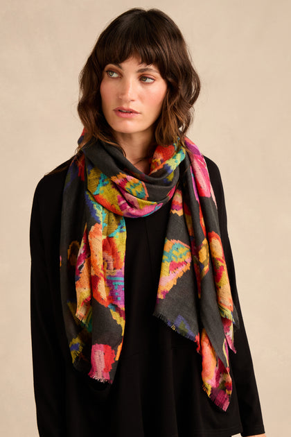 A woman stands against a plain beige background, looking slightly to the side, wearing a black top and the Pixelated Floral Wool Scarf.