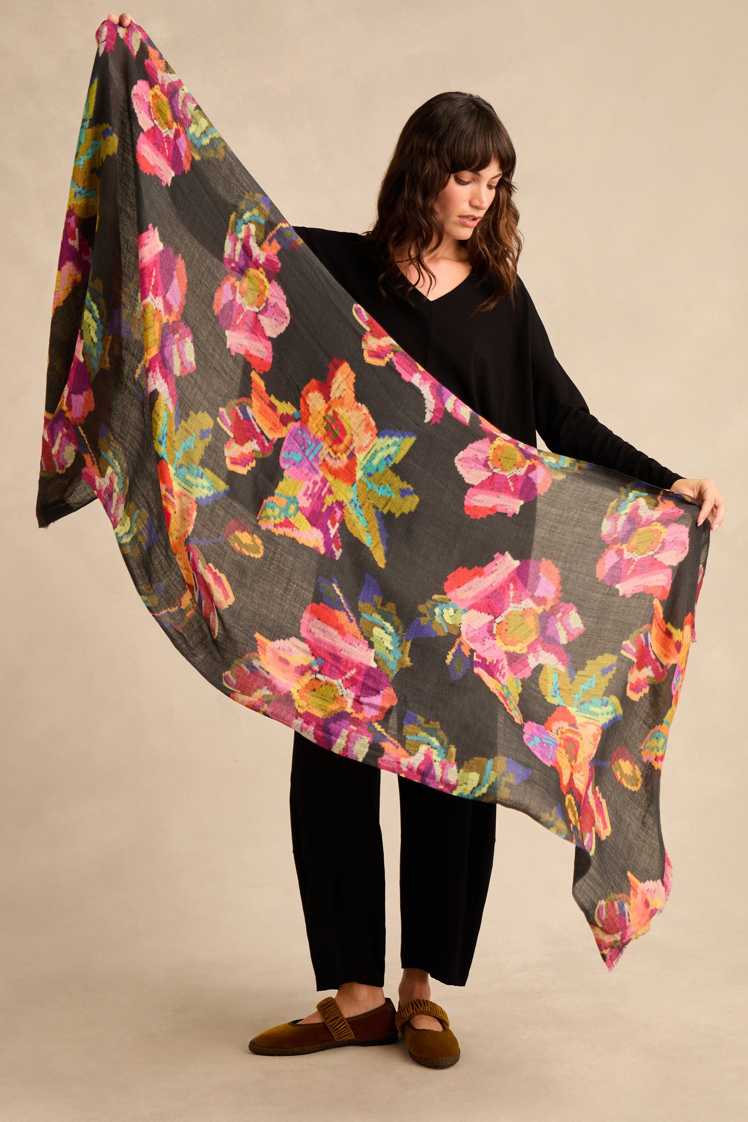 A person holds out the Pixelated Floral Wool Scarf, a large, sheer black accessory adorned with bright, colorful floral patterns.