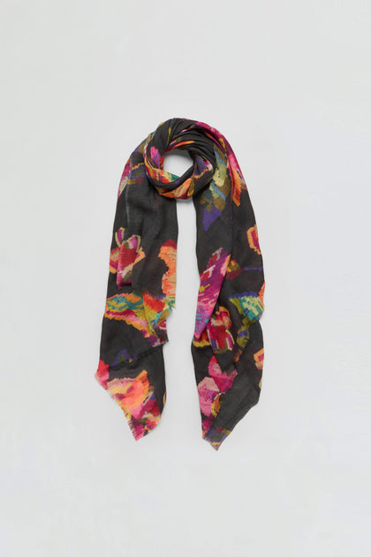 The Pixelated Floral Wool Scarf is a long, black wool-silk accessory showcasing a colorful, abstract botanical pattern in shades of pink, orange, red, and green, displayed flat on a white background.
