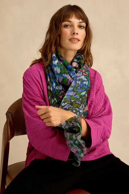 A woman sits on a chair against a neutral background, wearing a textured magenta jacket, black pants, and the Winter Garden Wool Scarf featuring a colorful botanical pattern.