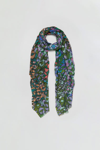 The Winter Garden Wool Scarf is a multicolored wool accessory with an abstract animal print in green, orange, purple, and blue hues, evoking the vibrancy of a winter garden and showcased against a simple light backdrop.