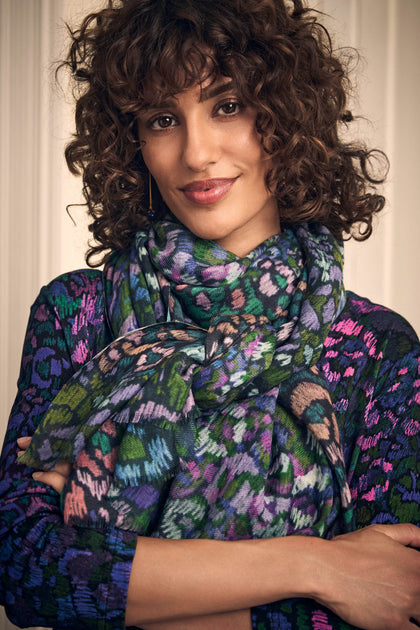 A woman with dark curly hair wears a Winter Garden Wool Scarf featuring a vibrant, multi-colored winter garden botanical pattern.
