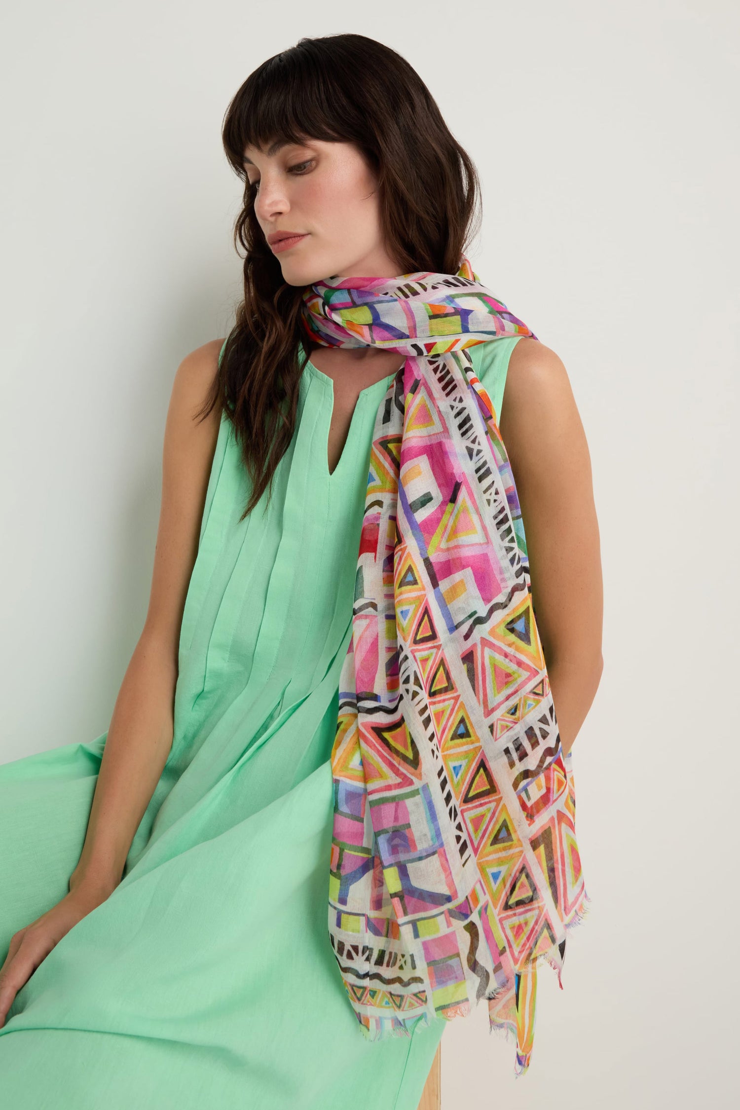 A woman in a sleeveless light green dress sits by a white wall, elegantly draping the Painted Geometric Scarf—crafted from luxe modal and silk with colorful geometric patterns—around her neck and shoulder.