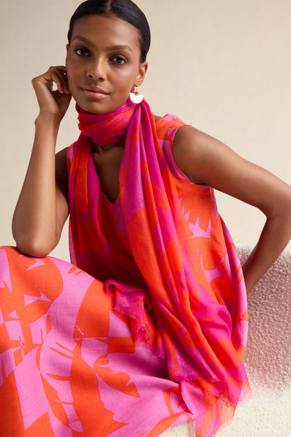 A woman wearing a sleeveless pink and orange patterned dress with the Palm Mirage Scarf sits on a textured chair, looking at the camera.