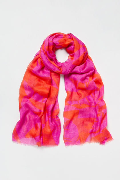 The Palm Mirage Scarf is a lightweight modal silk blend featuring an abstract tropical foliage pattern in vibrant pink and orange, shown loosely folded on a plain white background.
