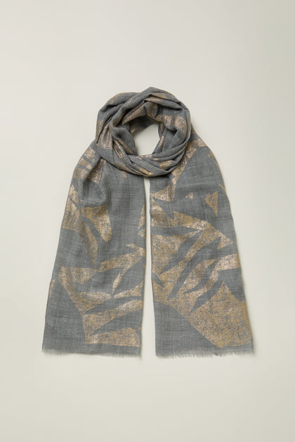 The Aurelian Cotton Scarf in grey displays abstract gold metallic patterns, lightly folded and laid flat on a light background.