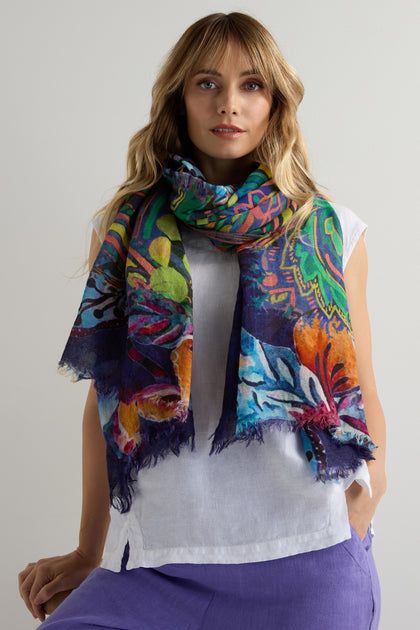 A woman in a white top and purple pants wears the Painted Paisley Wool Scarf, a lightweight accessory with a vibrant paisley design, draped around her neck against a plain light background.