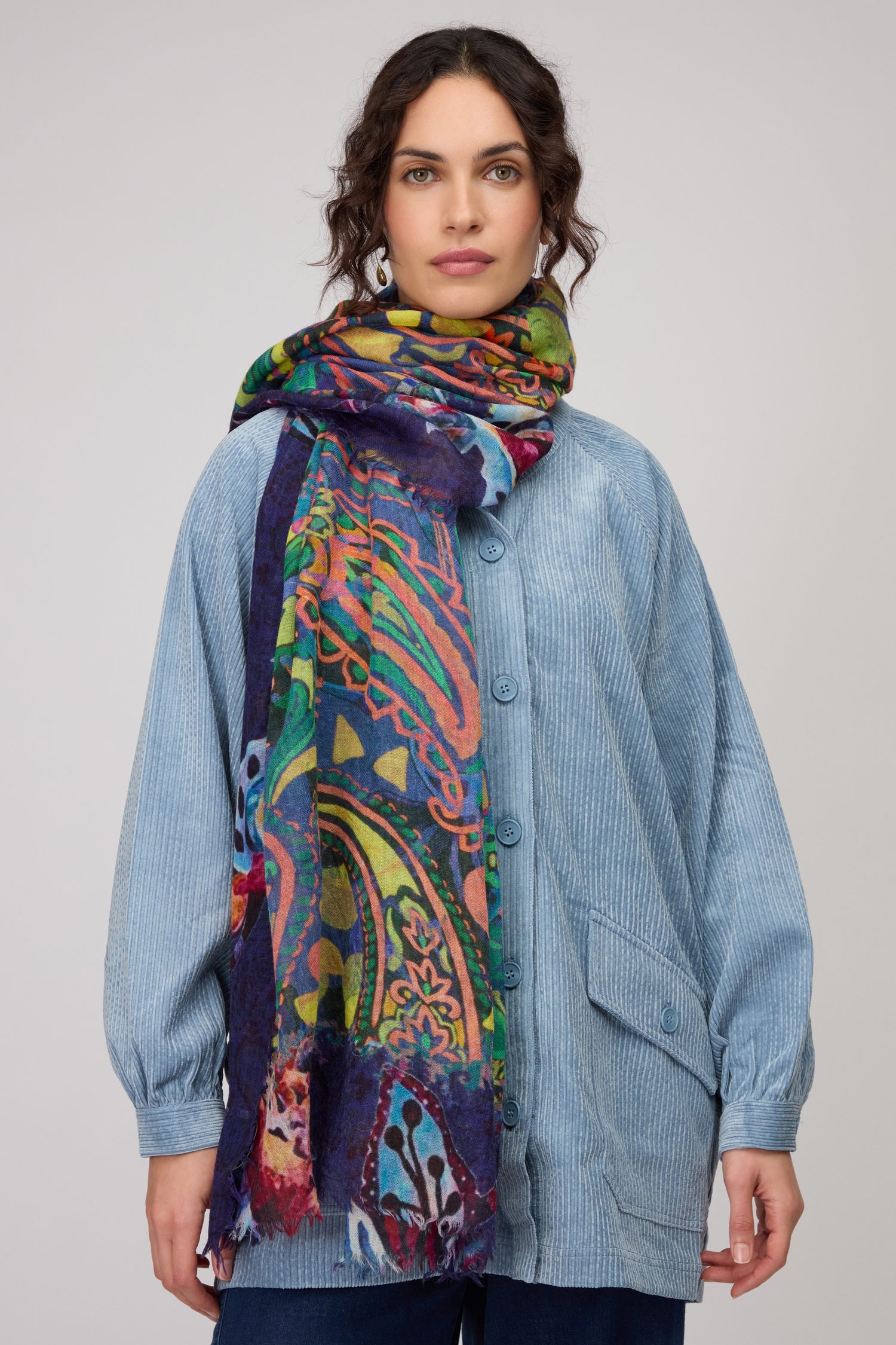 A woman faces the camera against a plain background, wearing a light blue corduroy jacket and a Painted Paisley Wool Scarf—a multicolored, lightweight wool accessory with a paisley design.