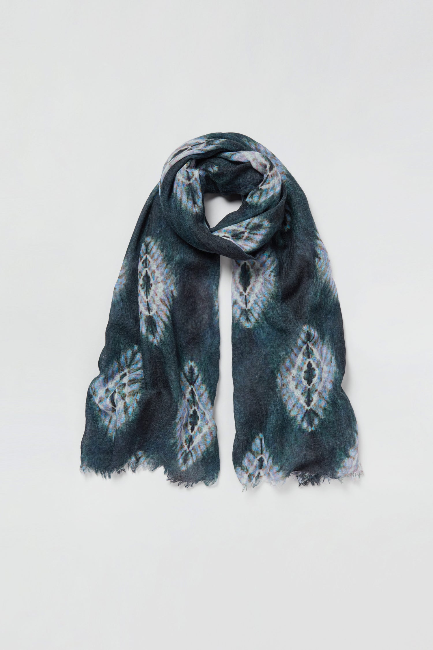 The Shibori Wool Scarf features a dark, tie-dye inspired pattern and fringed edges, creating a breathable and stylish accessory when laid flat on a white background.
