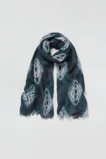 The Shibori Wool Scarf features a dark, tie-dye inspired pattern and fringed edges, creating a breathable and stylish accessory when laid flat on a white background.