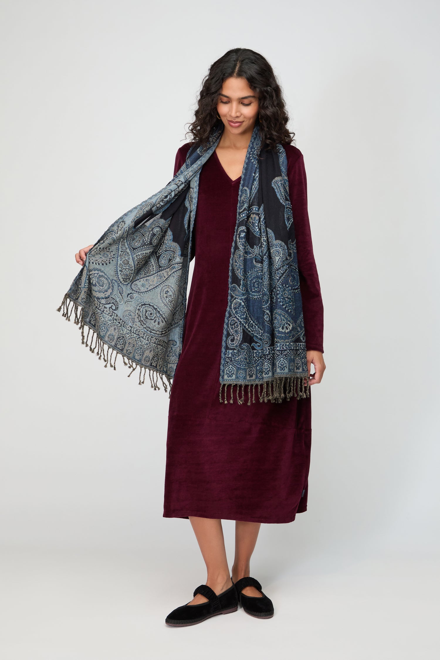 A woman stands against a plain background, wearing a long-sleeved burgundy dress, black shoes, and the Opulent Paisley Wool Scarf with fringe.
