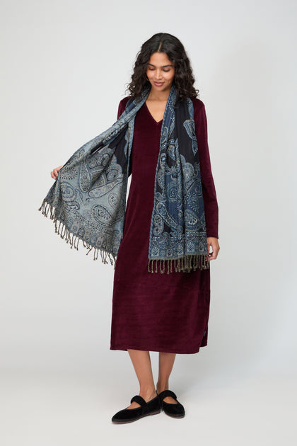 A woman stands against a plain background, wearing a long-sleeved burgundy dress, black shoes, and the Opulent Paisley Wool Scarf with fringe.
