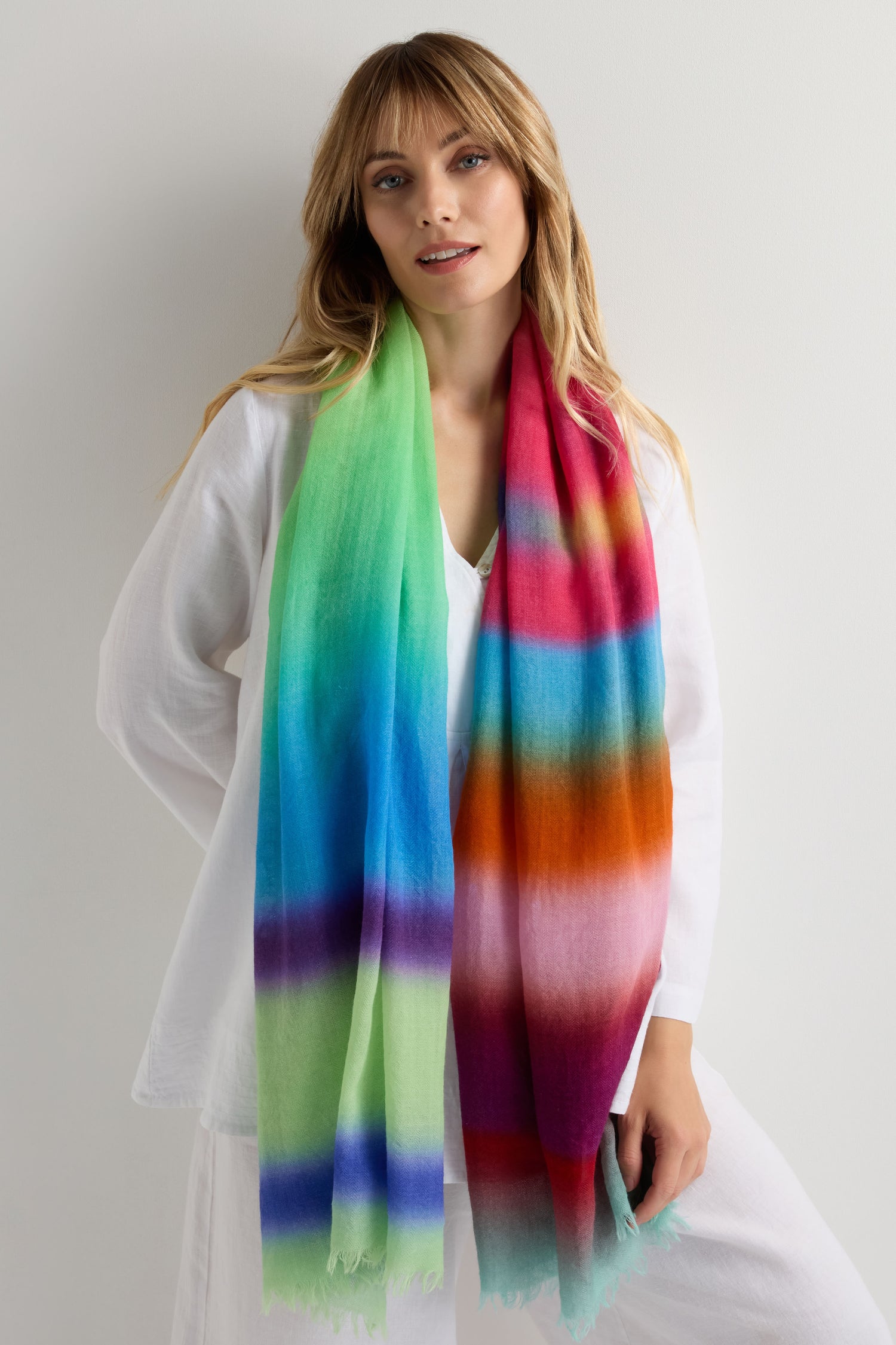 A woman in white poses against a plain backdrop, draped in the Horizon Stripe Wool Scarf—pure wool with an ombre of green, blue, purple, red, and orange stripes.