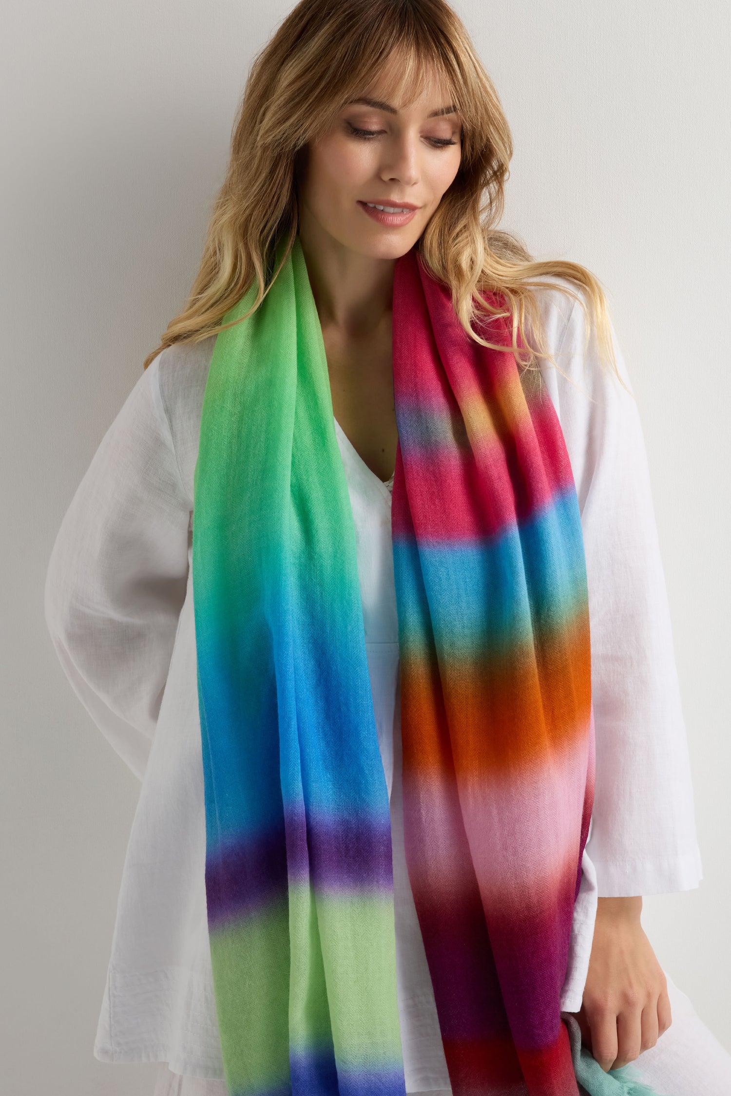 A woman in a white blouse wears the Horizon Stripe Wool Scarf, featuring a colorful ombre stripe design in green, blue, purple, red, and orange, as she stands against a neutral background.