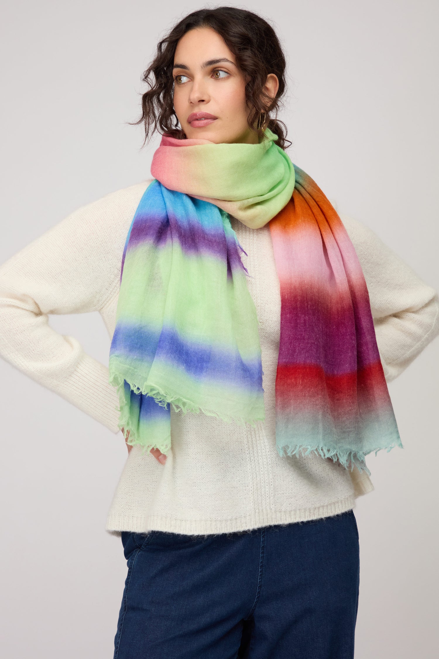 Woman wearing a cream sweater, blue pants, and the Horizon Stripe Wool Scarf—a large, lightweight wool scarf with an ombre design in green, blue, purple, red, and orange—stands against a plain background.