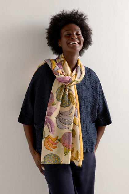 A person smiles in a textured black top and dark pants, accessorized with the vibrant Bird of Paradise Modal Scarf, whose colorful design evokes tropical landscapes. The scarf's ultra-soft modal fabric adds luxurious elegance to the outfit.