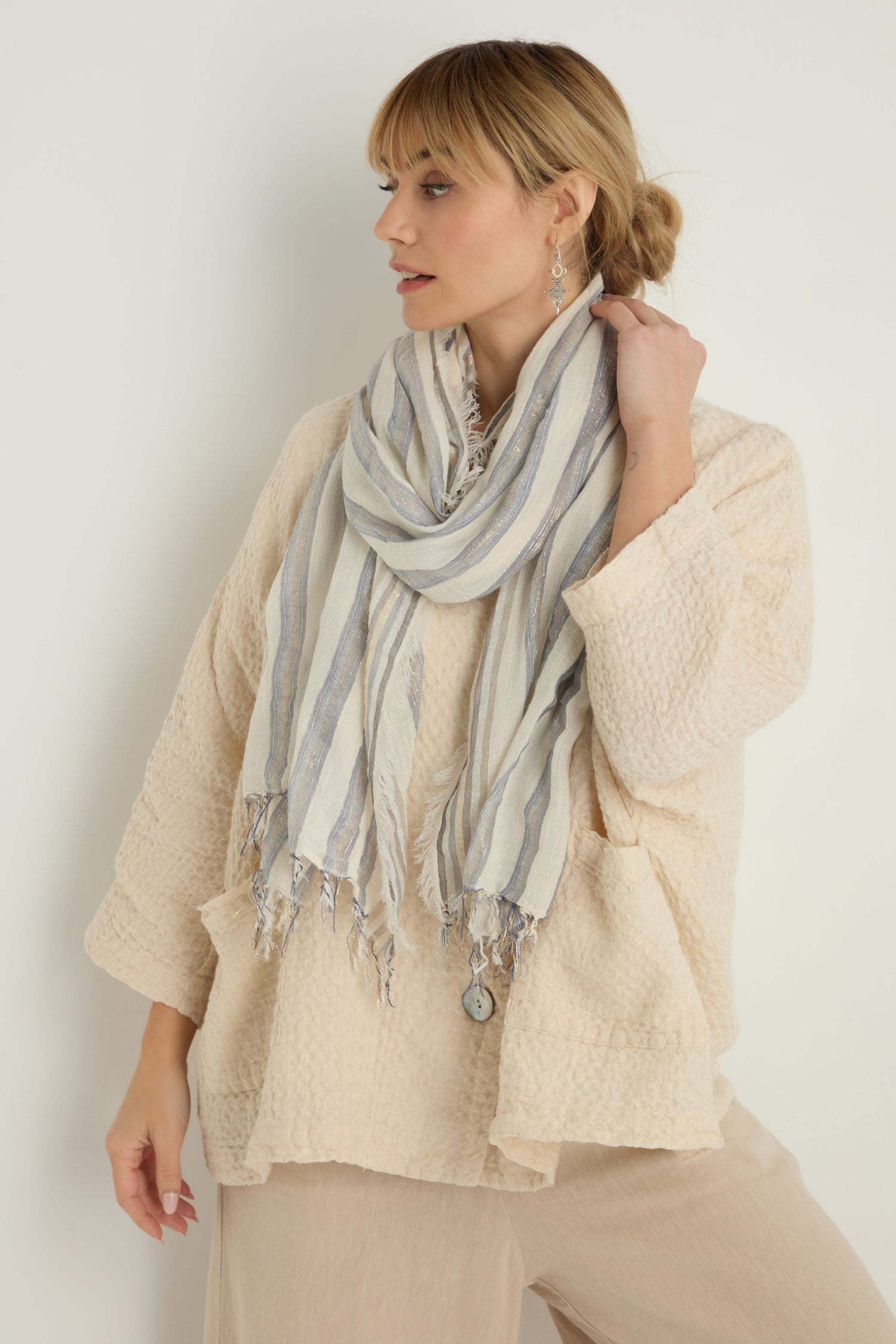 A woman in a beige textured top, matching pants, and the Lurex Stripe Cotton Blend Scarf looks to the left against a plain background.