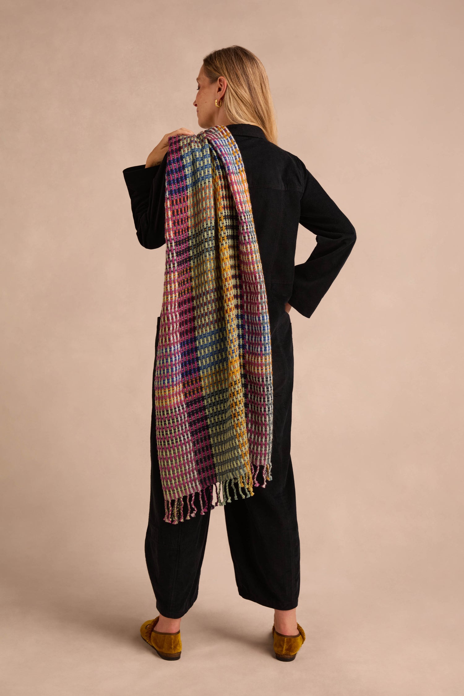 Wearing a black outfit and facing away, the person drapes the Checkerboard Scarf—a colorful wool-blend accessory—over their left shoulder, bringing lightweight warmth to a neutral backdrop.