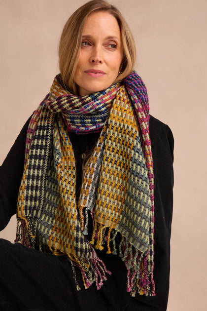 A woman wears the Checkerboard Scarf—an oversized, multicolored accessory—draped over a black coat. Seated with her arms crossed and gazing slightly to the side against a neutral background, she lets the lightweight wool-blend scarf add warmth and a vibrant pop to her ensemble.