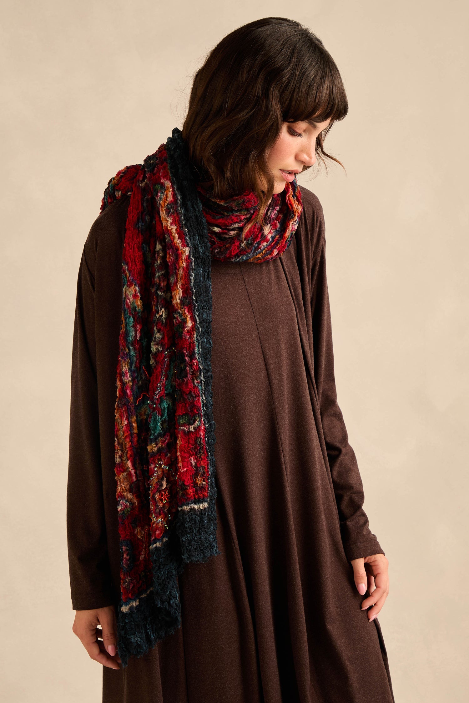 A person wearing a long brown dress and the Crushed Sequin Detail Wool Scarf in ruby, featuring subtle sequin accents and a textured finish, stands against a plain, light background, looking downward.