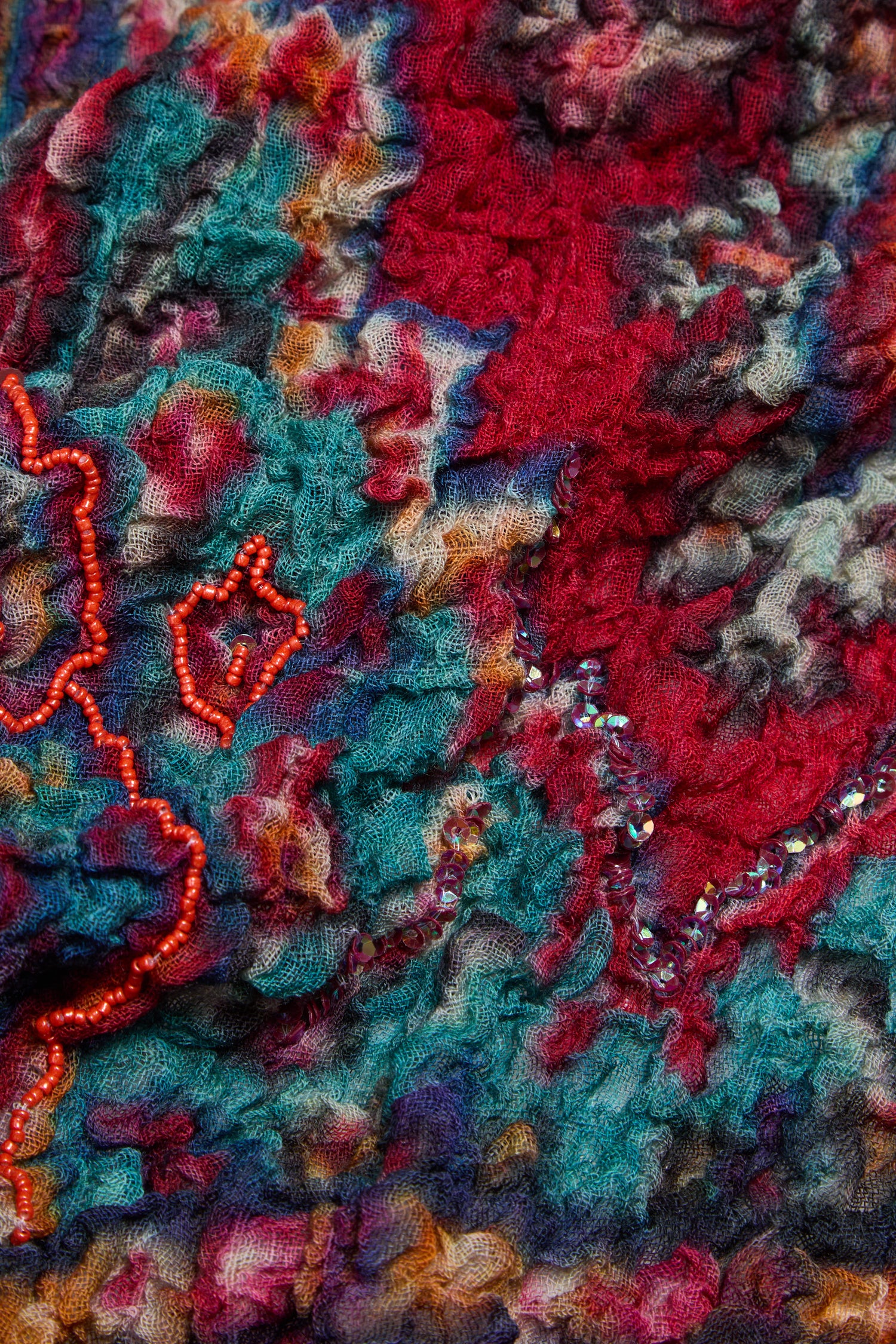 Close-up of the Crushed Sequin Detail Wool Scarf, showcasing its vibrant red, blue, and yellow patterned fabric with textured beaded embroidery. The crumpled, wool-like surface and subtle sequin accents add luxurious shimmer to this colorful design.